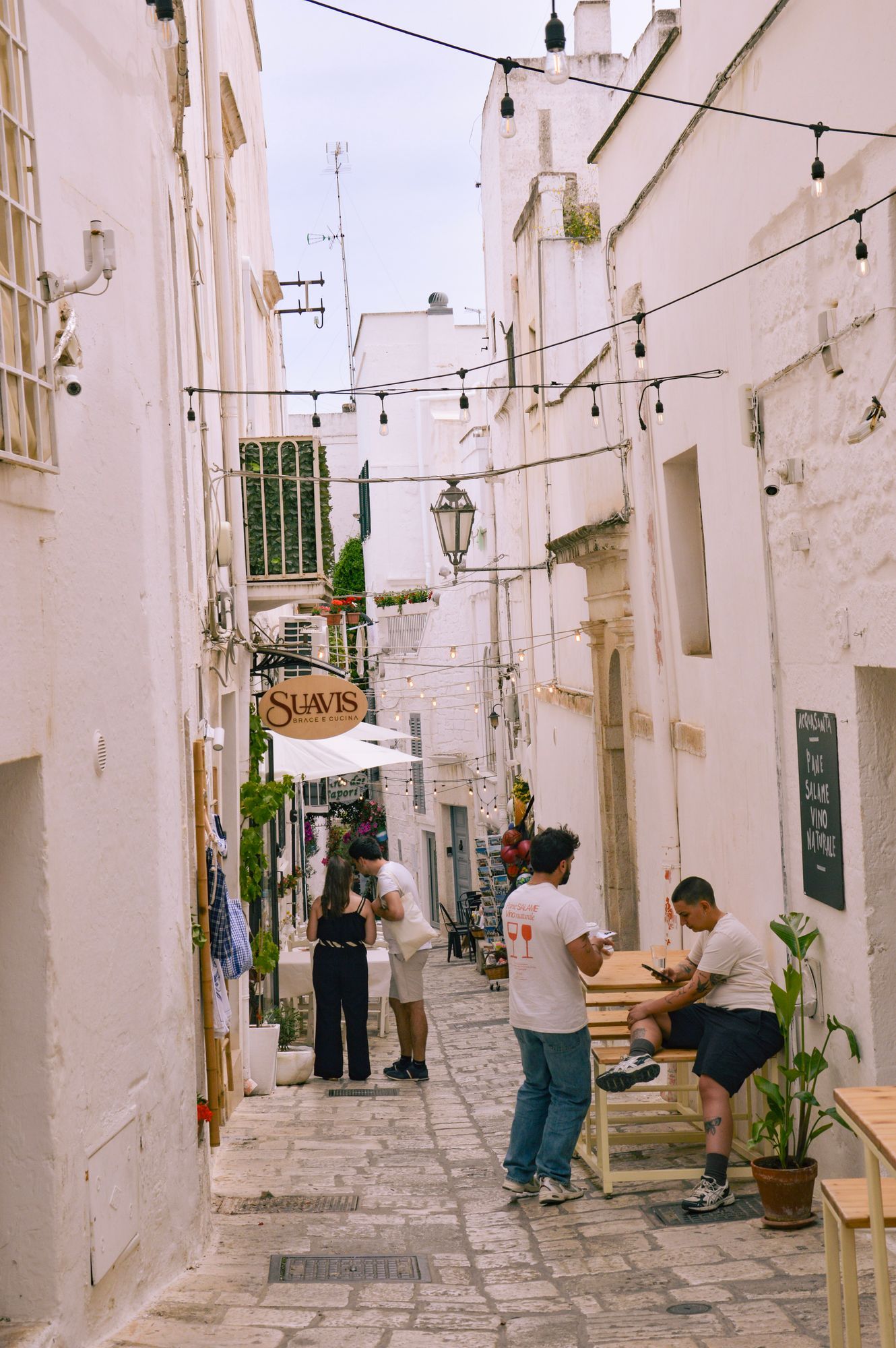 A white street in Ostuni in the late afternoon, with outdoor tables, lights and people.